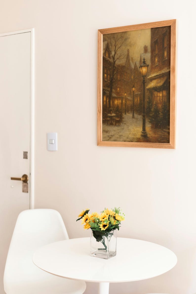 K&ouml;nnte beinhalten: Ein gerahmtes Gem&auml;lde einer verschneiten Stra&szlig;enszene mit einer Stra&szlig;enlaterne h&auml;ngt an einer Wand. Darunter steht ein kleiner wei&szlig;er Tisch mit einer klaren Glasvase mit Sonnenblumen. Ein wei&szlig;er Stuhl steht neben dem Tisch.