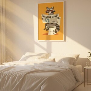 Op de afbeelding: Een ingelijste poster met een gele achtergrond met een wasbeer die een krant met de titel "THE DAILY DUMP" leest op een toilet. De poster hangt boven een bed met wit beddengoed.