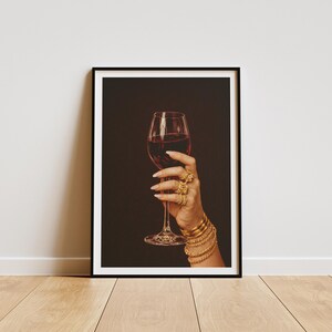 Peut inclure: Impression encadr&eacute;e d'une main tenant un verre de vin rouge. La main est orn&eacute;e de plusieurs bagues et bracelets dor&eacute;s. Le fond est brun fonc&eacute; et le cadre est noir. Le tout est pos&eacute; sur un sol en bois.