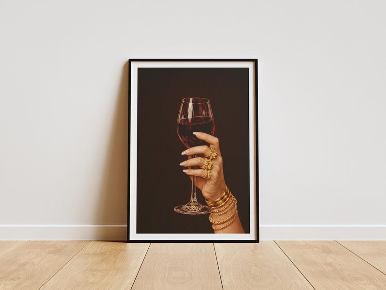 Peut inclure: Impression encadr&eacute;e d'une main tenant un verre de vin rouge. La main est orn&eacute;e de plusieurs bagues et bracelets dor&eacute;s. Le fond est brun fonc&eacute; et le cadre est noir. Le tout est pos&eacute; sur un sol en bois.
