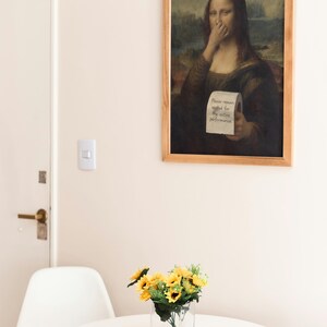 May include: A framed print of the Mona Lisa with a humorous twist. The artwork depicts the famous painting with a roll of toilet paper and the text "Please remain seated for the entire performance." A small vase of sunflowers sits on a white table.