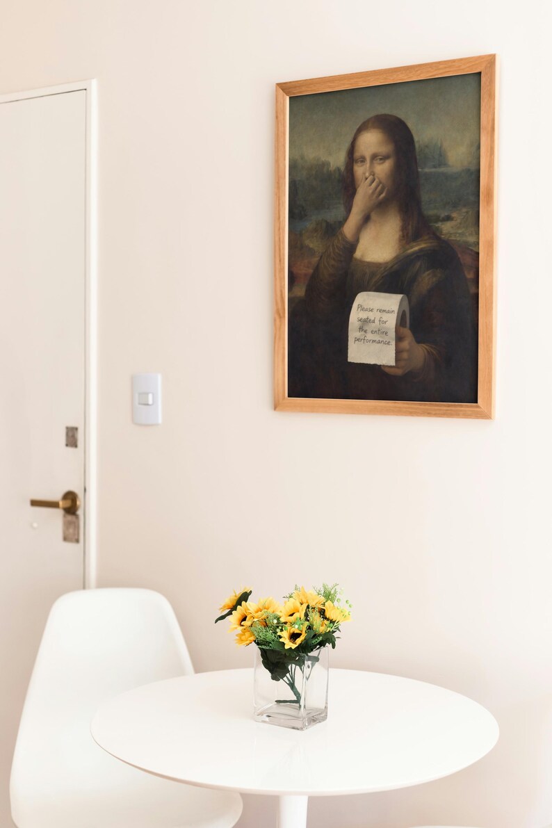 May include: A framed print of the Mona Lisa with a humorous twist. The artwork depicts the famous painting with a roll of toilet paper and the text "Please remain seated for the entire performance." A small vase of sunflowers sits on a white table.