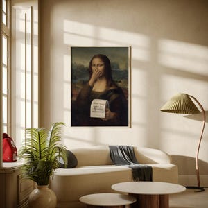 May include: A framed print of the Mona Lisa holding a tissue to her nose, with text that reads "Please remain seated for the entire performance." The print hangs above a cream-coloured sofa with a grey throw blanket. Two round coffee tables with wooden legs are in front of the sofa.
