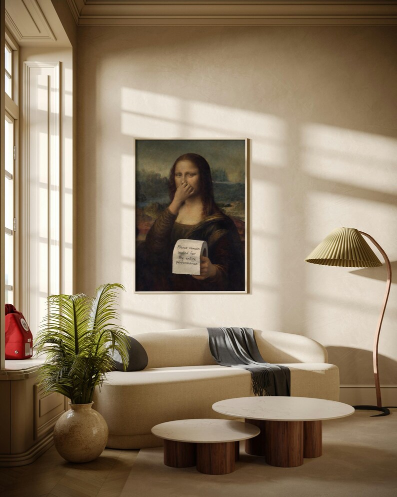May include: A framed print of the Mona Lisa holding a tissue to her nose, with text that reads "Please remain seated for the entire performance." The print hangs above a cream-coloured sofa with a grey throw blanket. Two round coffee tables with wooden legs are in front of the sofa.