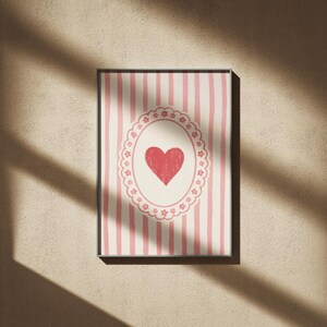 Op de afbeelding: Ingelijst kunstwerk met een rood hart in een decoratief ovaal op een gestreepte achtergrond. De strepen wisselen af tussen roze en wit. Het kunstwerk zit in een zwart frame en is op een beige muur gemonteerd.
