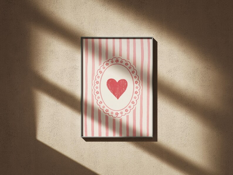 Op de afbeelding: Ingelijst kunstwerk met een rood hart in een decoratief ovaal op een gestreepte achtergrond. De strepen wisselen af tussen roze en wit. Het kunstwerk zit in een zwart frame en is op een beige muur gemonteerd.