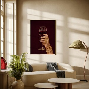 Peut inclure: Impression encadr&eacute;e d'une main tenant un verre de vin rouge. La main est orn&eacute;e de bagues et de bracelets dor&eacute;s. Le fond est bordeaux fonc&eacute;. L'impression est dans un salon moderne avec un canap&eacute; et des tables basses.