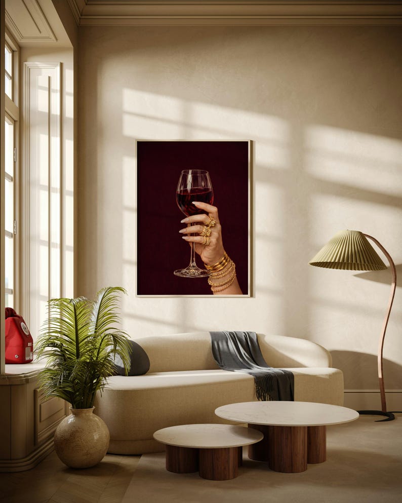 Peut inclure: Impression encadr&eacute;e d'une main tenant un verre de vin rouge. La main est orn&eacute;e de bagues et de bracelets dor&eacute;s. Le fond est bordeaux fonc&eacute;. L'impression est dans un salon moderne avec un canap&eacute; et des tables basses.