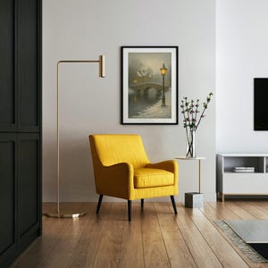 May include: A framed print of a bridge over water with a lamppost, in a room with a yellow armchair, a gold floor lamp, and a white side table. The room has hardwood floors and a black cabinet.