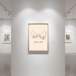 Puede incluir: Impresi&oacute;n art&iacute;stica enmarcada con una ilustraci&oacute;n minimalista de dos manos entrelazando los me&ntilde;iques, con un coraz&oacute;n y las palabras "you & me" en cursiva. La obra de arte se exhibe en una galer&iacute;a con otras piezas enmarcadas.