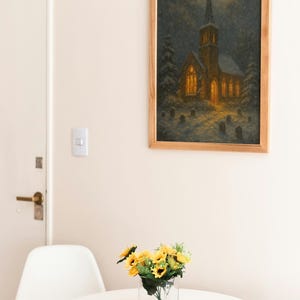Peut inclure: Un tableau encadr&eacute; d'une &eacute;glise la nuit, avec un clocher et des fen&ecirc;tres &eacute;clair&eacute;es, est accroch&eacute; au mur. En dessous, une petite table blanche accueille un vase en verre transparent avec des tournesols jaunes.