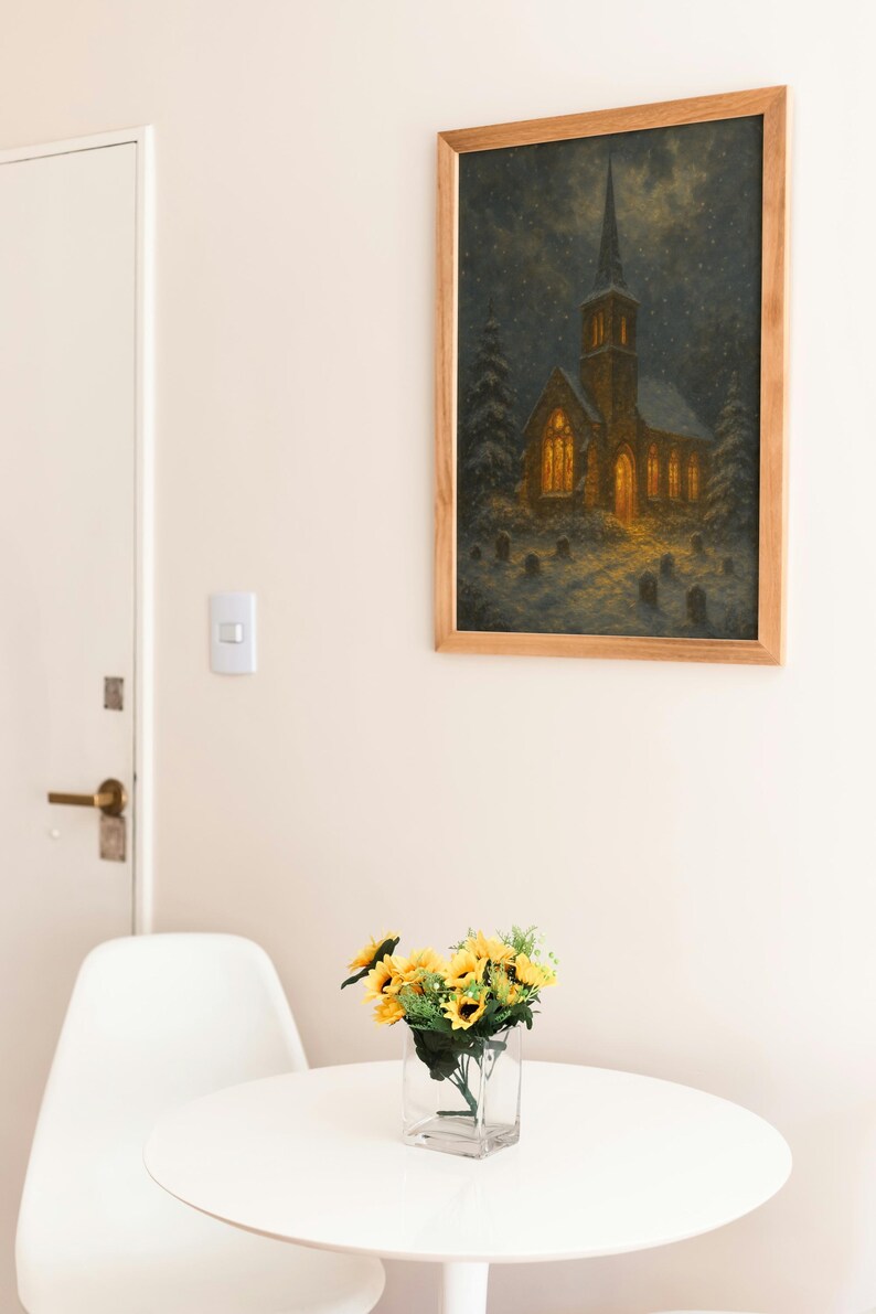 Peut inclure: Un tableau encadr&eacute; d'une &eacute;glise la nuit, avec un clocher et des fen&ecirc;tres &eacute;clair&eacute;es, est accroch&eacute; au mur. En dessous, une petite table blanche accueille un vase en verre transparent avec des tournesols jaunes.
