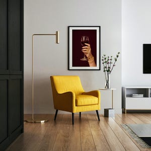 Peut inclure: Un salon moderne avec un fauteuil jaune, une lampe de sol dor&eacute;e et une impression encadr&eacute;e d'une main tenant un verre de vin rouge. La pi&egrave;ce a des planchers de bois franc et une table d'appoint blanche avec un vase de fleurs.