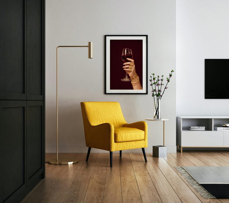 Peut inclure: Un salon moderne avec un fauteuil jaune, une lampe de sol dor&eacute;e et une impression encadr&eacute;e d'une main tenant un verre de vin rouge. La pi&egrave;ce a des planchers de bois franc et une table d'appoint blanche avec un vase de fleurs.