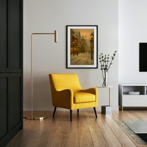 May include: A framed winter street scene painting hangs above a yellow armchair with black legs. A gold floor lamp stands to the left, and a white side table with a vase of flowers is to the right. The room has hardwood floors.