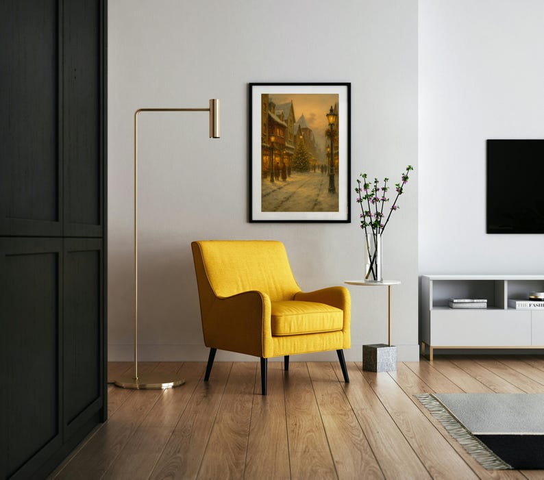 May include: A framed winter street scene painting hangs above a yellow armchair with black legs. A gold floor lamp stands to the left, and a white side table with a vase of flowers is to the right. The room has hardwood floors.