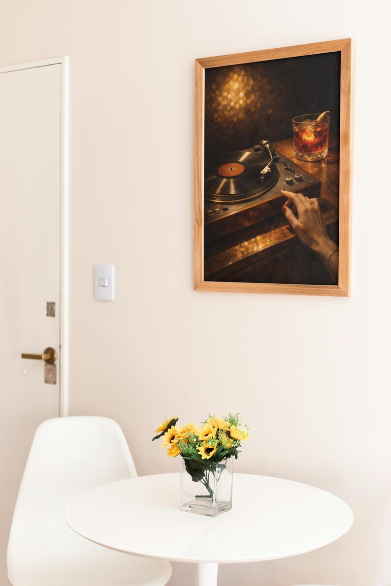 May include: A framed print showing a vintage record player and a cocktail glass. The artwork is set against a warm, blurred background. A small vase of sunflowers sits on a white table. The room has a minimalist design.