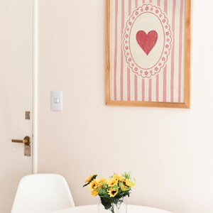 Op de afbeelding: Een ingelijst kunstwerk met een rood hartontwerp op een roze gestreepte achtergrond hangt aan een muur. Daaronder staat een kleine witte tafel met een heldere glazen vaas met gele zonnebloemen. Een witte stoel staat naast de tafel.