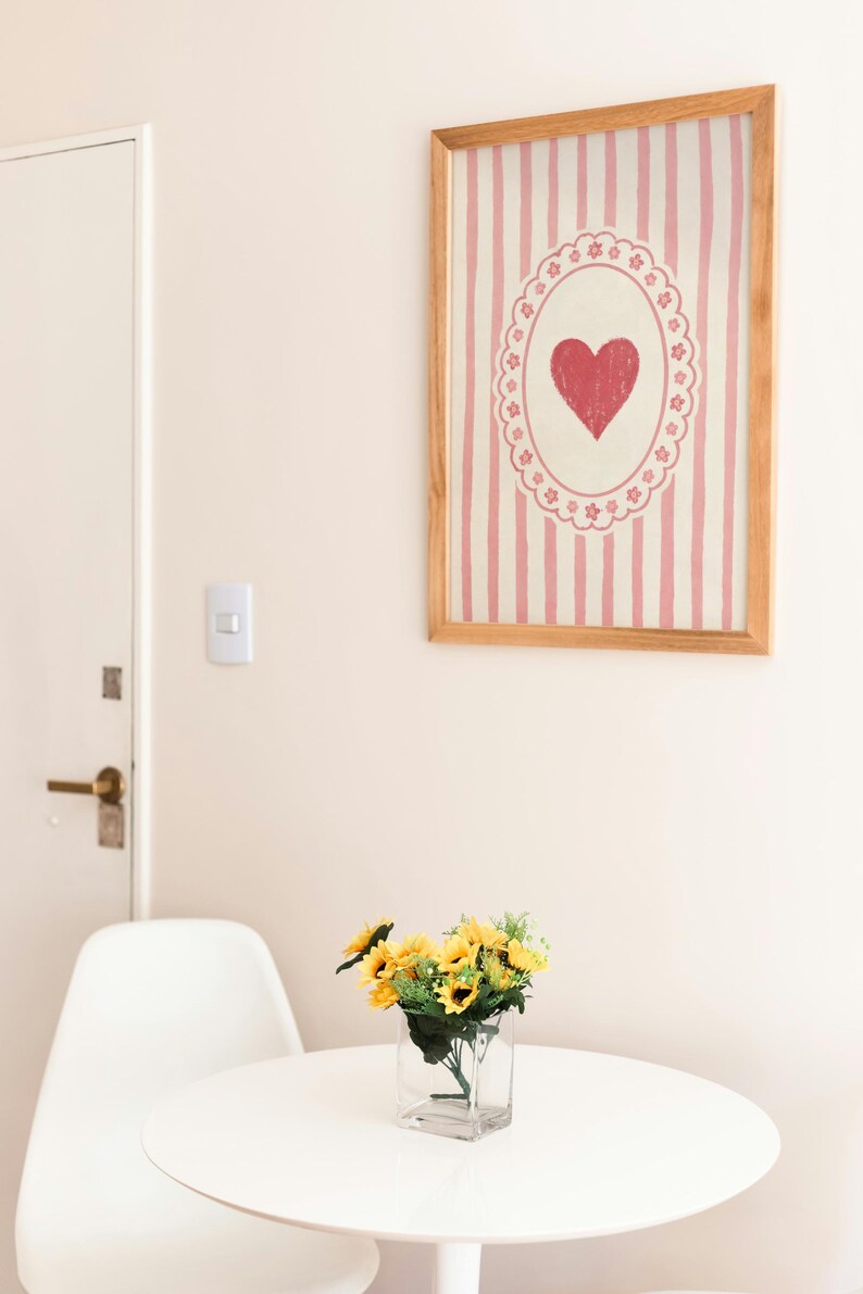Op de afbeelding: Een ingelijst kunstwerk met een rood hartontwerp op een roze gestreepte achtergrond hangt aan een muur. Daaronder staat een kleine witte tafel met een heldere glazen vaas met gele zonnebloemen. Een witte stoel staat naast de tafel.