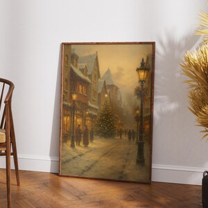 May include: Framed vintage painting of a snowy European street scene at dusk. The artwork depicts a Christmas tree, street lamps, and figures walking along a snow-covered road. The painting has a warm, inviting color palette.