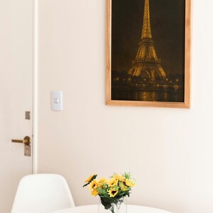 May include: A framed print of the Eiffel Tower at night hangs on a wall. A small white table holds a clear glass vase with yellow sunflowers. A white chair is next to the table.