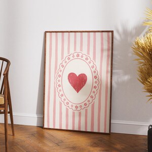 Op de afbeelding: Een ingelijst kunstwerk met een rood hart in een decoratief ovaal op een gestreepte achtergrond. Het kunstwerk heeft een houten lijst en leunt tegen een witte muur. Het kleurenpalet omvat roze, rood en cr&egrave;me.