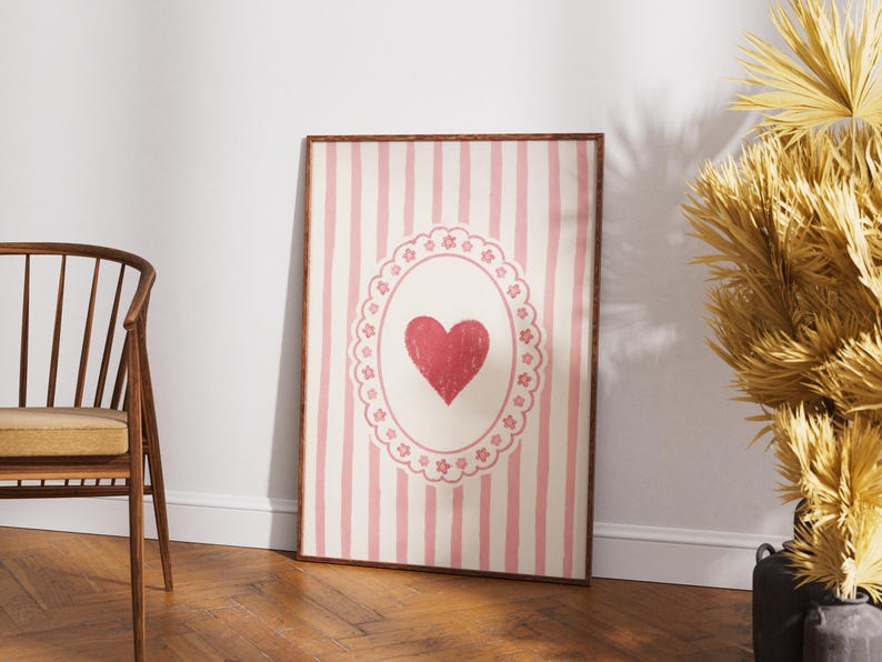 Op de afbeelding: Een ingelijst kunstwerk met een rood hart in een decoratief ovaal op een gestreepte achtergrond. Het kunstwerk heeft een houten lijst en leunt tegen een witte muur. Het kleurenpalet omvat roze, rood en cr&egrave;me.