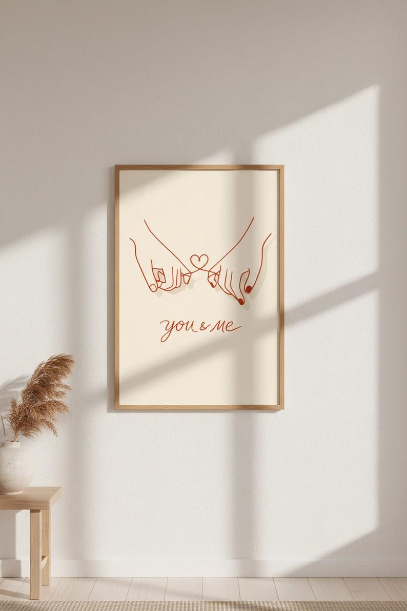 Puede incluir: Un cuadro enmarcado con un dibujo lineal minimalista de dos manos entrelazadas formando un coraz&oacute;n. La obra de arte es en rojo sobre un fondo beige, con las palabras "you & me" escritas debajo.