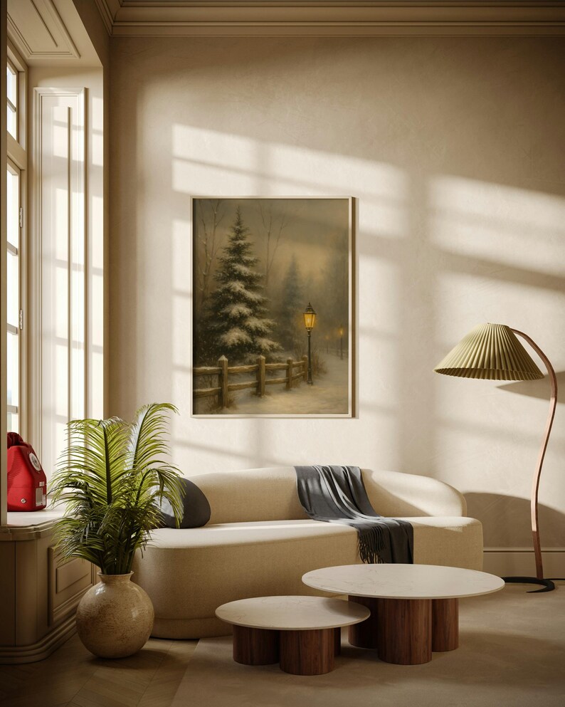 May include: A cozy living room scene featuring a framed winter landscape painting above a cream-colored sofa. Two round coffee tables with wooden bases and marble tops sit in front of the sofa. A green leafy plant in a beige vase adds a touch of nature.