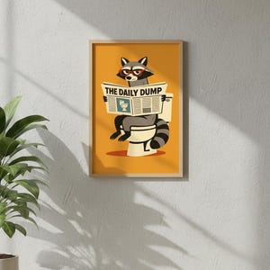 Op de afbeelding: Een ingelijste kunstprint met een cartoon wasbeer met een bril die een krant leest met de titel "THE DAILY DUMP" terwijl hij op een toilet zit. De achtergrond is effen oranje.