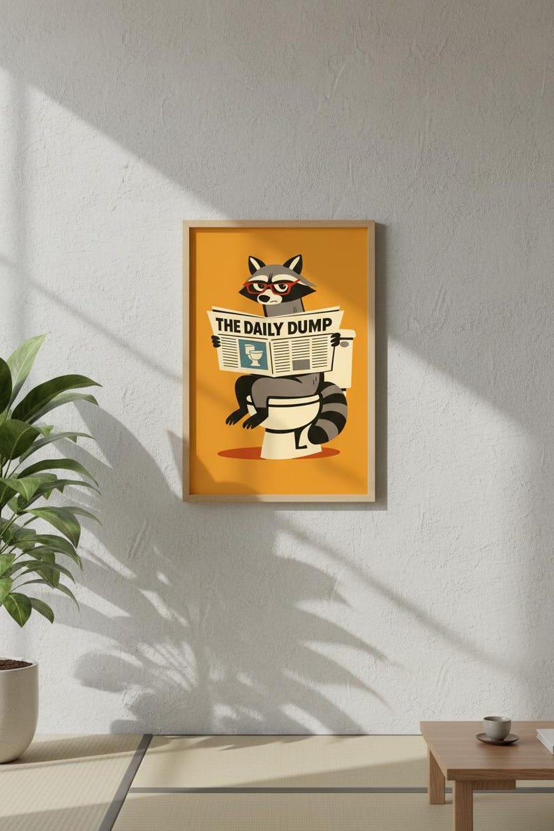 Op de afbeelding: Een ingelijste kunstprint met een cartoon wasbeer met een bril die een krant leest met de titel "THE DAILY DUMP" terwijl hij op een toilet zit. De achtergrond is effen oranje.