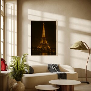 May include: A framed print of the Eiffel Tower at night hangs above a cream-colored sofa. Two round coffee tables with wooden bases and marble tops sit in front of the sofa. A green leafy plant in a beige vase is on the left.