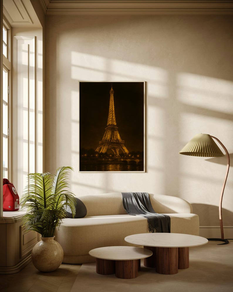 May include: A framed print of the Eiffel Tower at night hangs above a cream-colored sofa. Two round coffee tables with wooden bases and marble tops sit in front of the sofa. A green leafy plant in a beige vase is on the left.