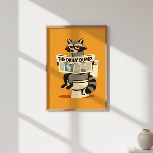 Op de afbeelding: Een ingelijste print met een wasbeer met een bril die een krant leest met de titel "THE DAILY DUMP" terwijl hij op een toilet zit. Het kunstwerk heeft een gele achtergrond en een humoristisch thema.