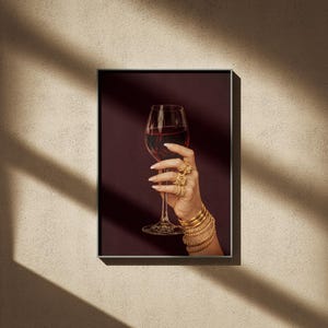 Peut inclure: Impression d'art encadr&eacute;e d'une main tenant un verre de vin rouge. La main est orn&eacute;e de bracelets et de bagues dor&eacute;es. Le fond est bordeaux fonc&eacute;, cr&eacute;ant une ambiance &eacute;l&eacute;gante.