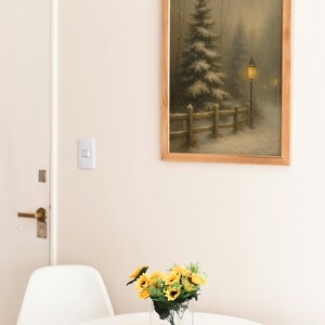May include: A framed landscape painting depicts a snowy scene with a tall evergreen tree, a wooden fence, and a lamplit path. A small bouquet of sunflowers sits in a clear glass vase on a white round table. A white chair is next to the table.