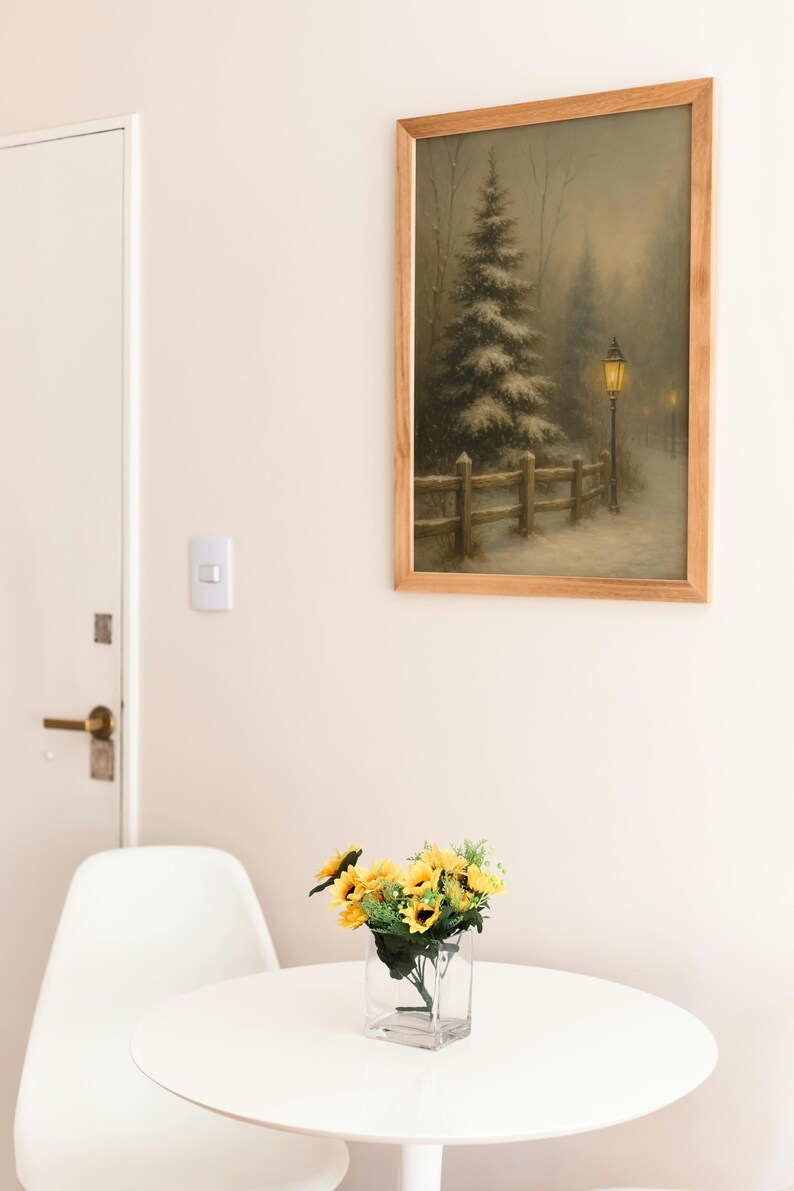 May include: A framed landscape painting depicts a snowy scene with a tall evergreen tree, a wooden fence, and a lamplit path. A small bouquet of sunflowers sits in a clear glass vase on a white round table. A white chair is next to the table.