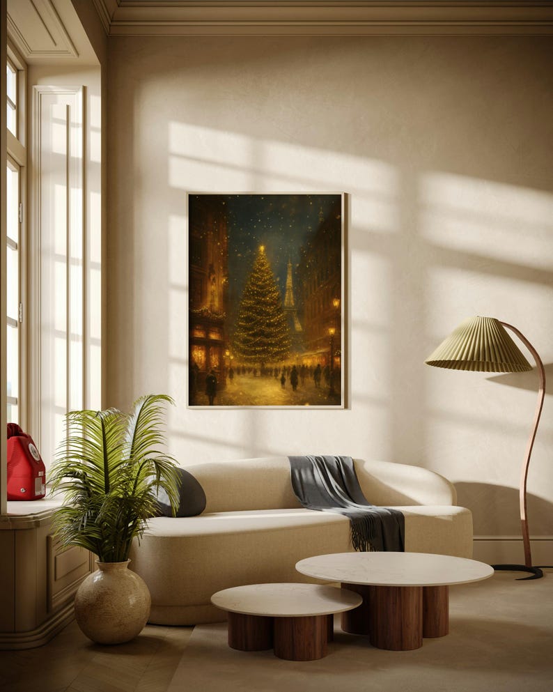 May include: A framed print of a Christmas scene in a city street, featuring a decorated tree and the Eiffel Tower. The print hangs above a cream-colored sofa with a gray throw, and two round coffee tables with wooden legs. A green plant in a beige vase sits nearby.