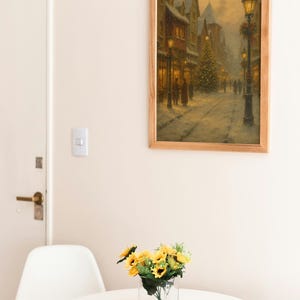 May include: A framed print of a snowy street scene with a Christmas tree and lit buildings. The artwork is in a light wood frame, hanging above a small white table with a vase of sunflowers. A white chair is next to the table.