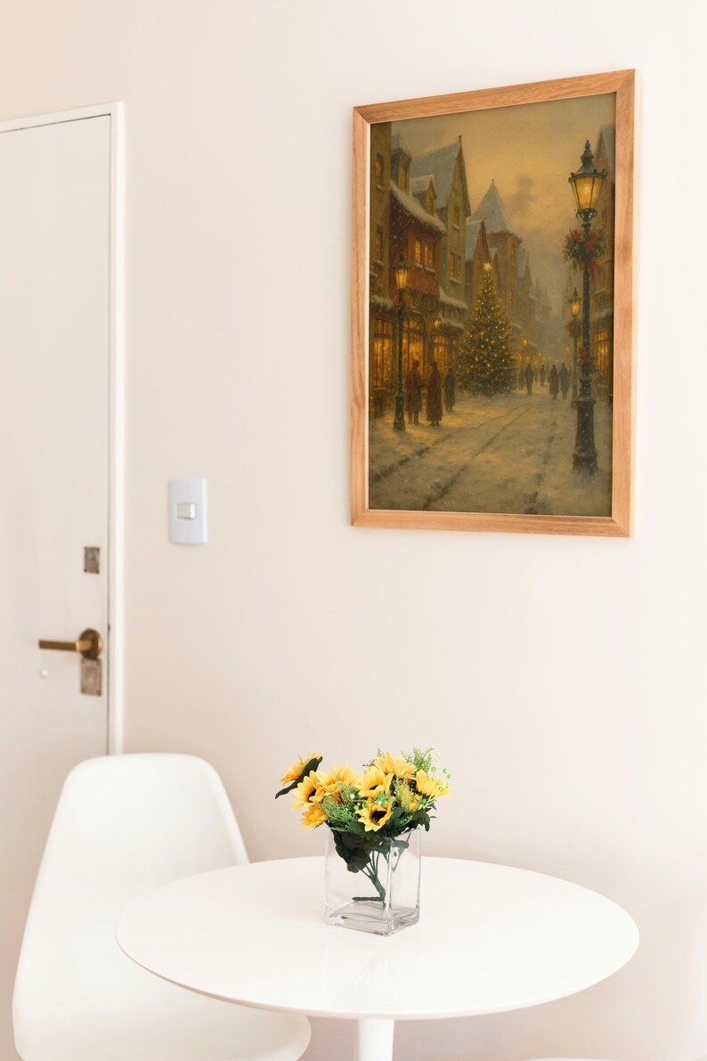 May include: A framed print of a snowy street scene with a Christmas tree and lit buildings. The artwork is in a light wood frame, hanging above a small white table with a vase of sunflowers. A white chair is next to the table.