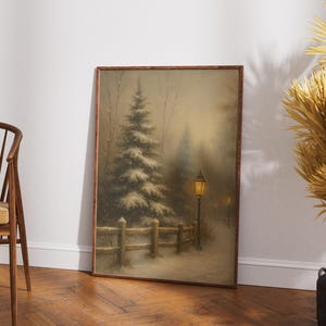May include: Framed winter landscape art print featuring a snow-covered scene with a tall evergreen tree, a wooden fence, and a lit lamppost. The artwork has a warm, muted color palette and is displayed on a wooden floor.