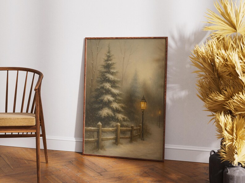 May include: Framed winter landscape art print featuring a snow-covered scene with a tall evergreen tree, a wooden fence, and a lit lamppost. The artwork has a warm, muted color palette and is displayed on a wooden floor.