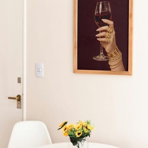 Peut inclure: Une impression encadr&eacute;e montre une main tenant un verre de vin rouge, orn&eacute;e de bijoux dor&eacute;s. L'impression est dans un cadre en bois, accroch&eacute;e &agrave; un mur cr&egrave;me. Une petite table avec un vase de tournesols est au premier plan.