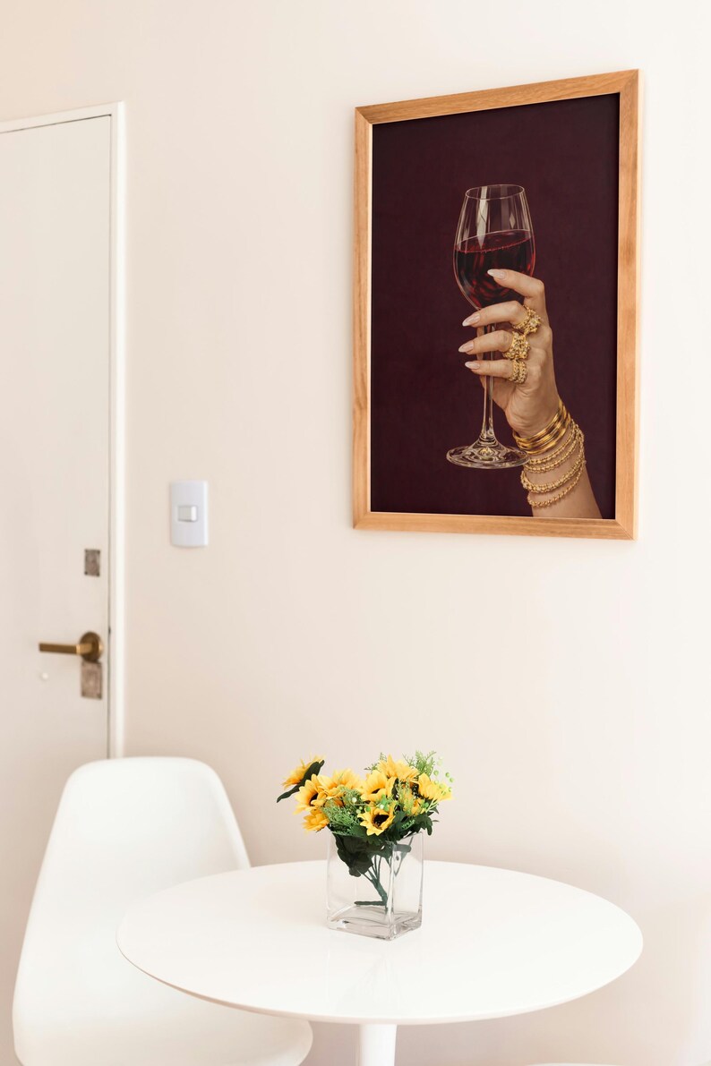 Peut inclure: Une impression encadr&eacute;e montre une main tenant un verre de vin rouge, orn&eacute;e de bijoux dor&eacute;s. L'impression est dans un cadre en bois, accroch&eacute;e &agrave; un mur cr&egrave;me. Une petite table avec un vase de tournesols est au premier plan.