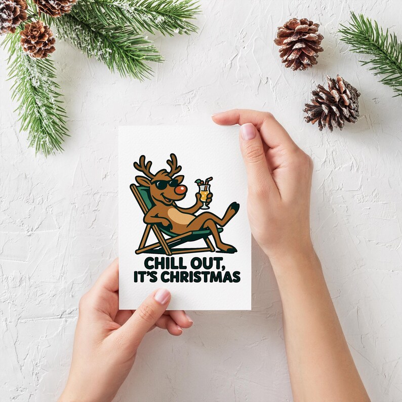 Pu&ograve; includere: Un biglietto di auguri bianco con un cartone animato di una renna con occhiali da sole e che si rilassa su una sedia a sdraio, con in mano un cocktail. Il biglietto recita "CHILL OUT, IT'S CHRISTMAS" in testo nero e verde. Il biglietto &egrave; tenuto in mano.