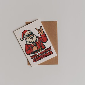 K&ouml;nnte beinhalten: Eine Weihnachtskarte mit einem Cartoon-Weihnachtsmann mit Sonnenbrille und roter Lederjacke, der ein Mikrofon h&auml;lt und eine Rock'n'Roll-Geste macht. Die Karte tr&auml;gt die Aufschrift "Have a Rockin' Christmas" und liegt auf einem braunen Umschlag.