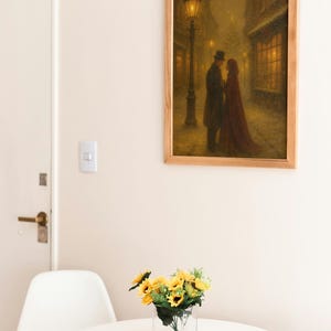 May include: A framed print shows a couple in a snowy street, lit by a gas lamp. The artwork, in warm tones, hangs above a white table with a vase of sunflowers. A white chair and a door are also visible.