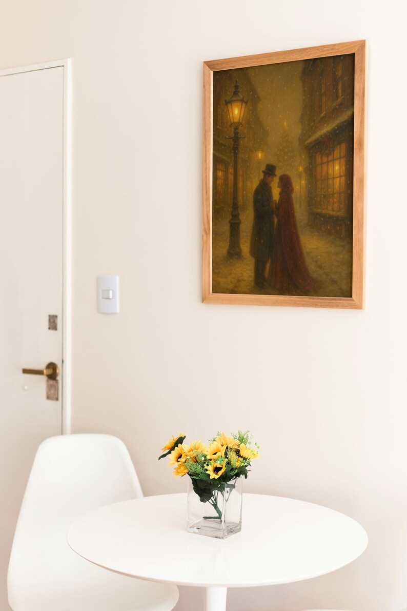 May include: A framed print shows a couple in a snowy street, lit by a gas lamp. The artwork, in warm tones, hangs above a white table with a vase of sunflowers. A white chair and a door are also visible.