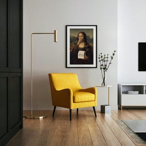 May include: A contemporary living room with a framed print of the Mona Lisa, humorously altered. A yellow armchair with black legs is positioned below the artwork. A gold floor lamp and a small side table with a vase of flowers are also present.