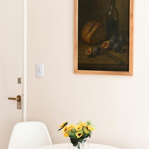 Pode incluir: Uma pintura emoldurada de natureza morta retrata um p&atilde;o, figos, uma faca e uma garrafa de vinho. Um pequeno vaso de girass&oacute;is est&aacute; em uma mesa branca em frente a uma cadeira branca.